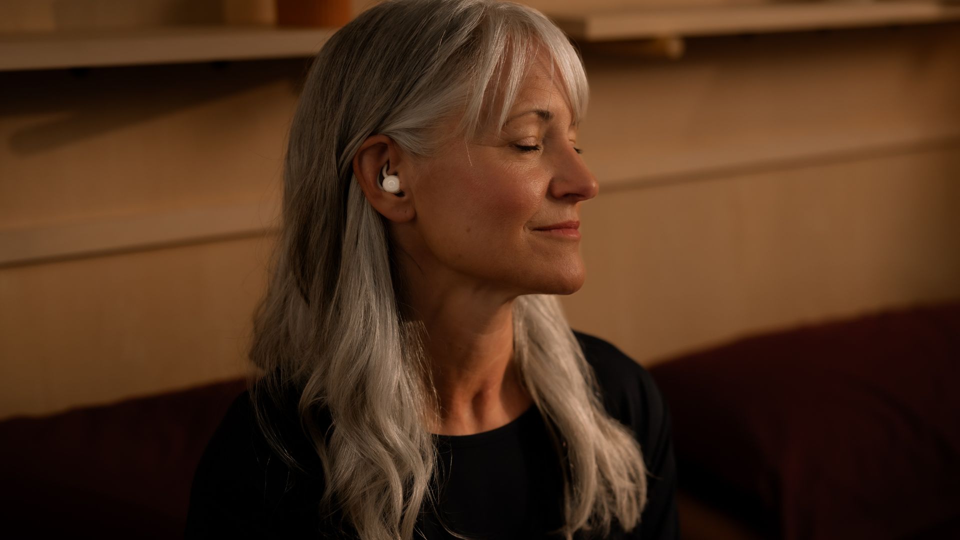 The image shows a woman with grey hair sitting down with her eyes closed while wearing a pair of white Ozlo Sleepbuds 2 while
