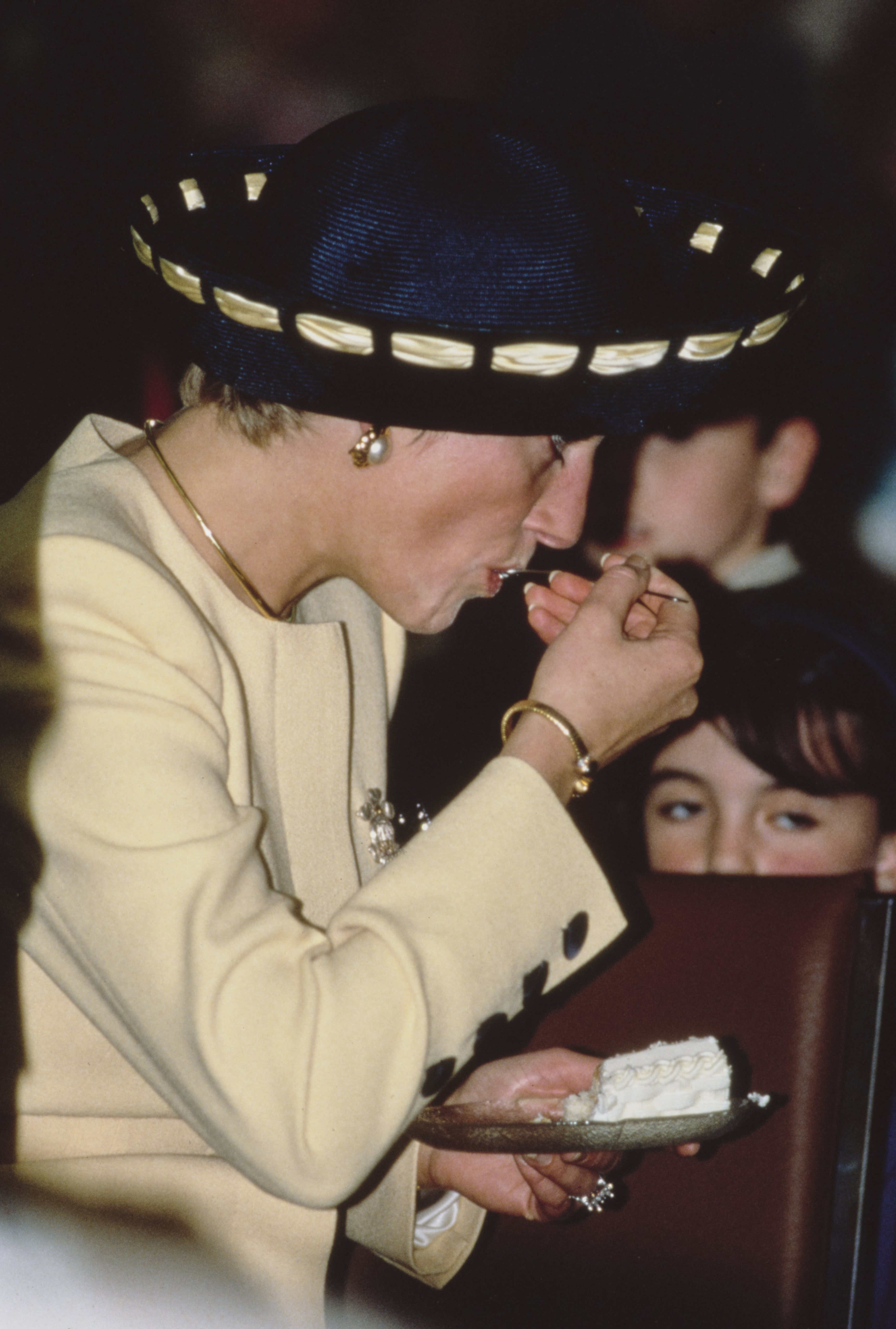 Princess Diana eating a piece of cake in a yellow suit and blue hat