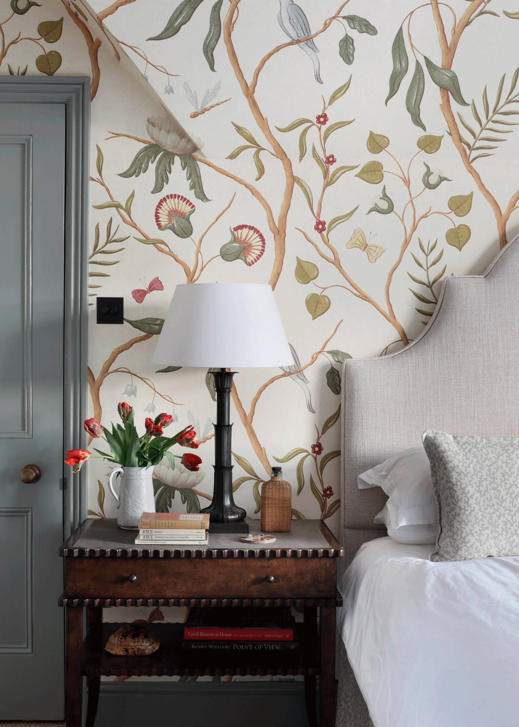 A bedroom with leafy wallpaper and a nightstand with a black table lamp, a base of tulips, a stack of books, and a bottle