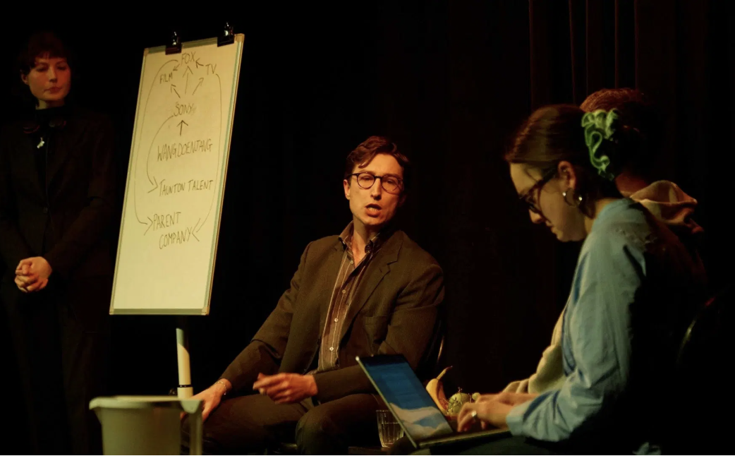 a production image form the play showing a man talking to a woman in a scene in a workplace