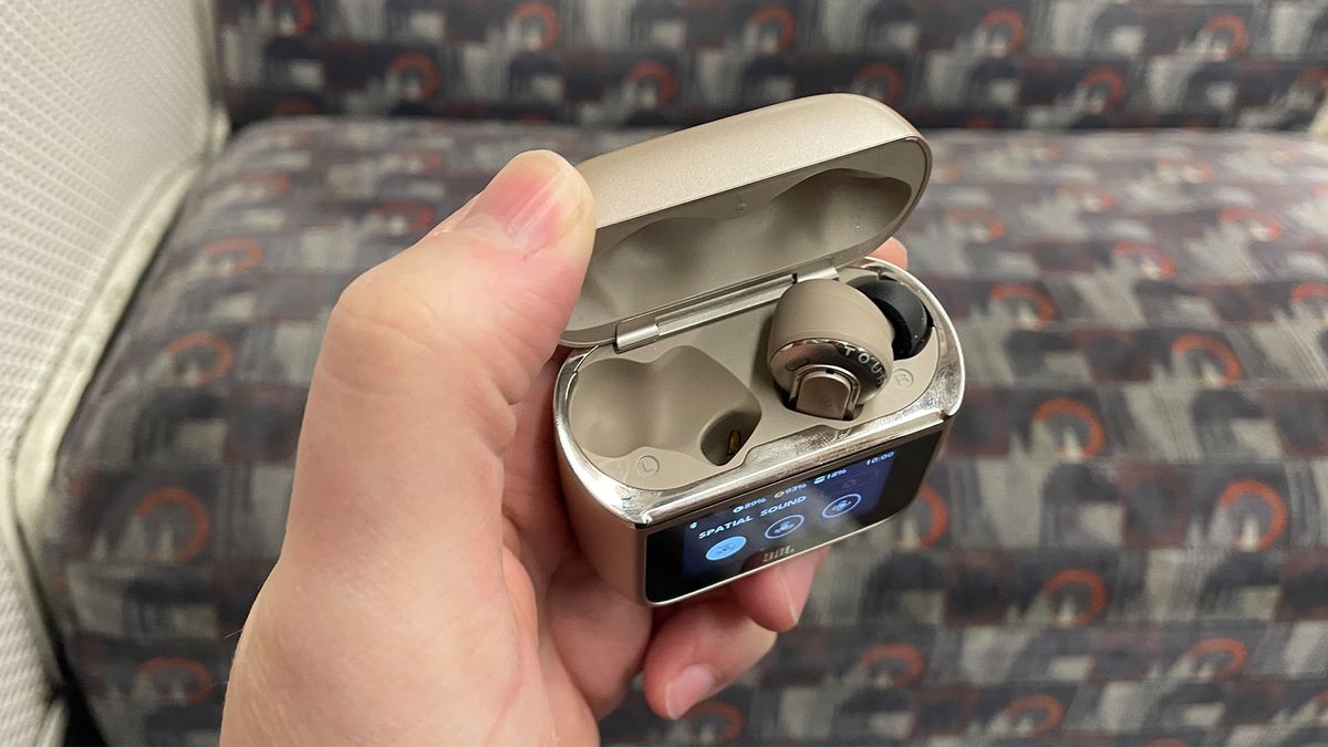 A hand holding the case for some JBL Tour Pro 3 earbuds. The lid is open and one of the buds can be seen inside, with the other compartment empty. The screen on the front of the case is just about visible, but you can't make out what it is displaying.