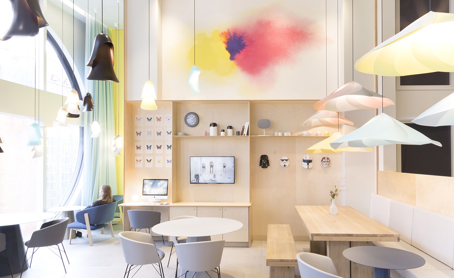 The bright reception area consists of high ceilings, light wooden furniture in combination with chairs in various shades of gray, and round tables with white tops.