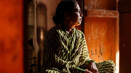 A woman in a green and white striped sari sits crosslegged in a wooden room, looking to the right of the camera