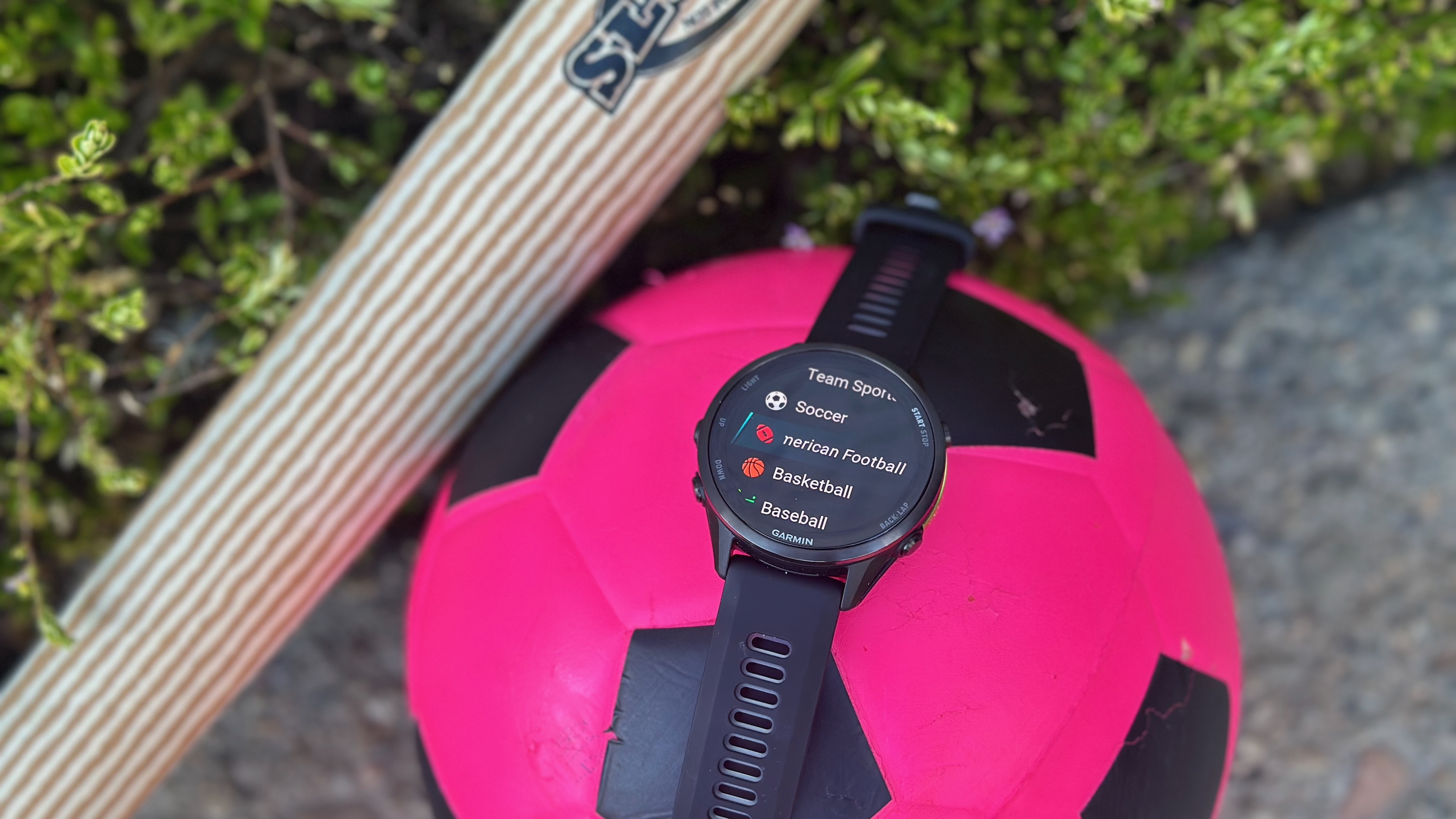 A Garmin Forerunner 970 sitting atop a pink soccer ball on pavement, with a kid's baseball bat next to it leaned against a bush.