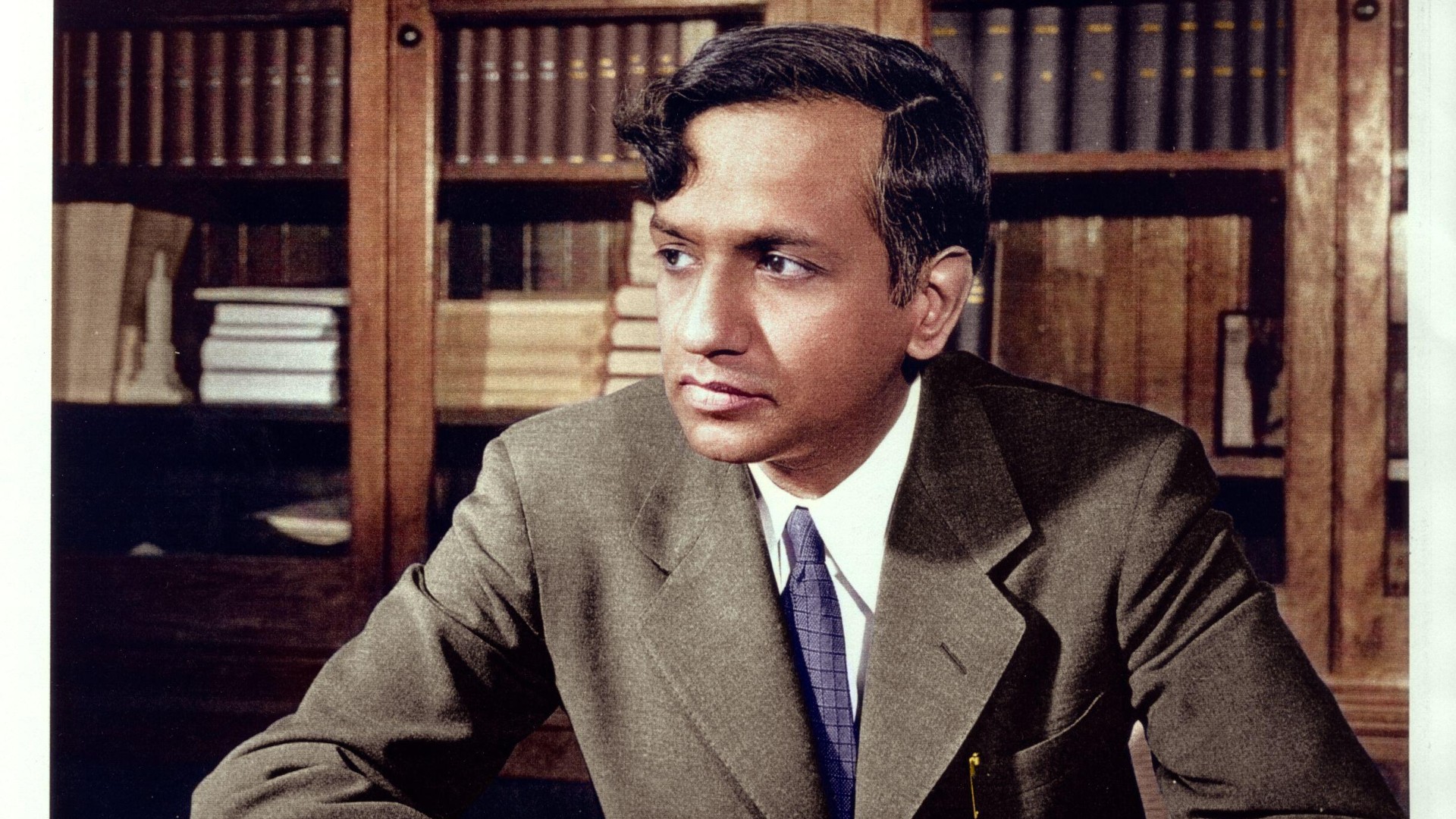 Photo of Subrahmanyan Chandrasekhar sitting at a desk with a bookshelf behind him.