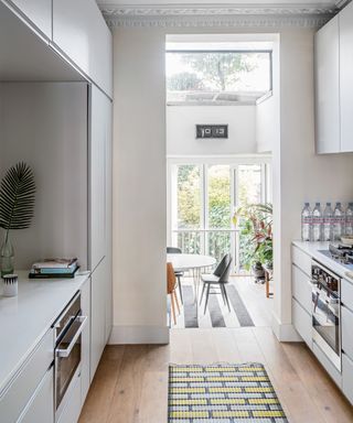 A white galley kitchen with handleless doors leading onto a dining area with large windows.