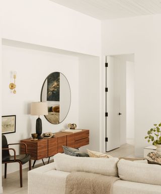 Round mirror above a wooden console at the head of a neutral living room with white couches