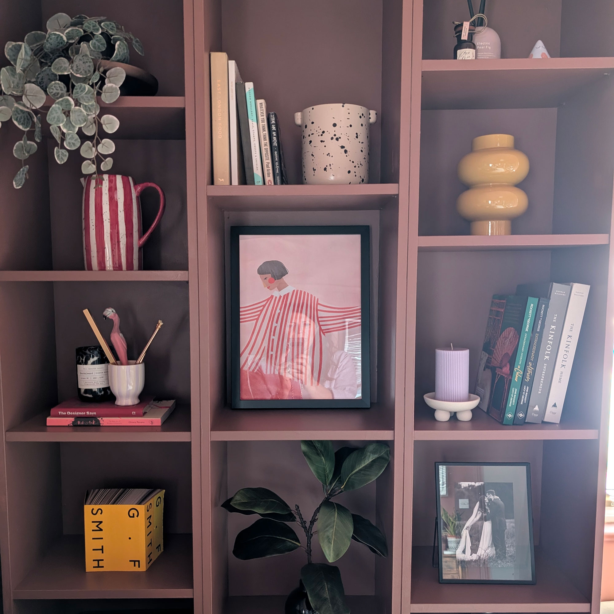 Dusky pink painted shelving unit with artwork, books, plants and vases