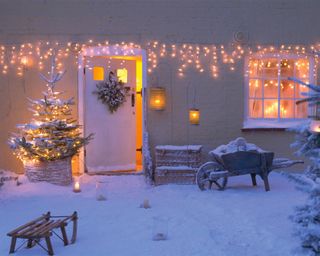 A snowy garden with twinkling string lights and a wreath lit front door