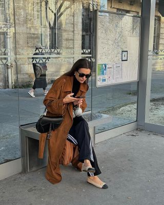 Woman wears Adidas track pants, a Dior saddle bag, Chanel flats and a brown trench coat while texting on the street.