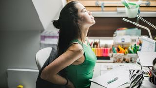 A woman feels her back as she sits at a desk. 