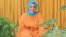 Nadiya Hussain wears an orange dress and turquoise hijab, sitting among plants and an orange fabric backdrop