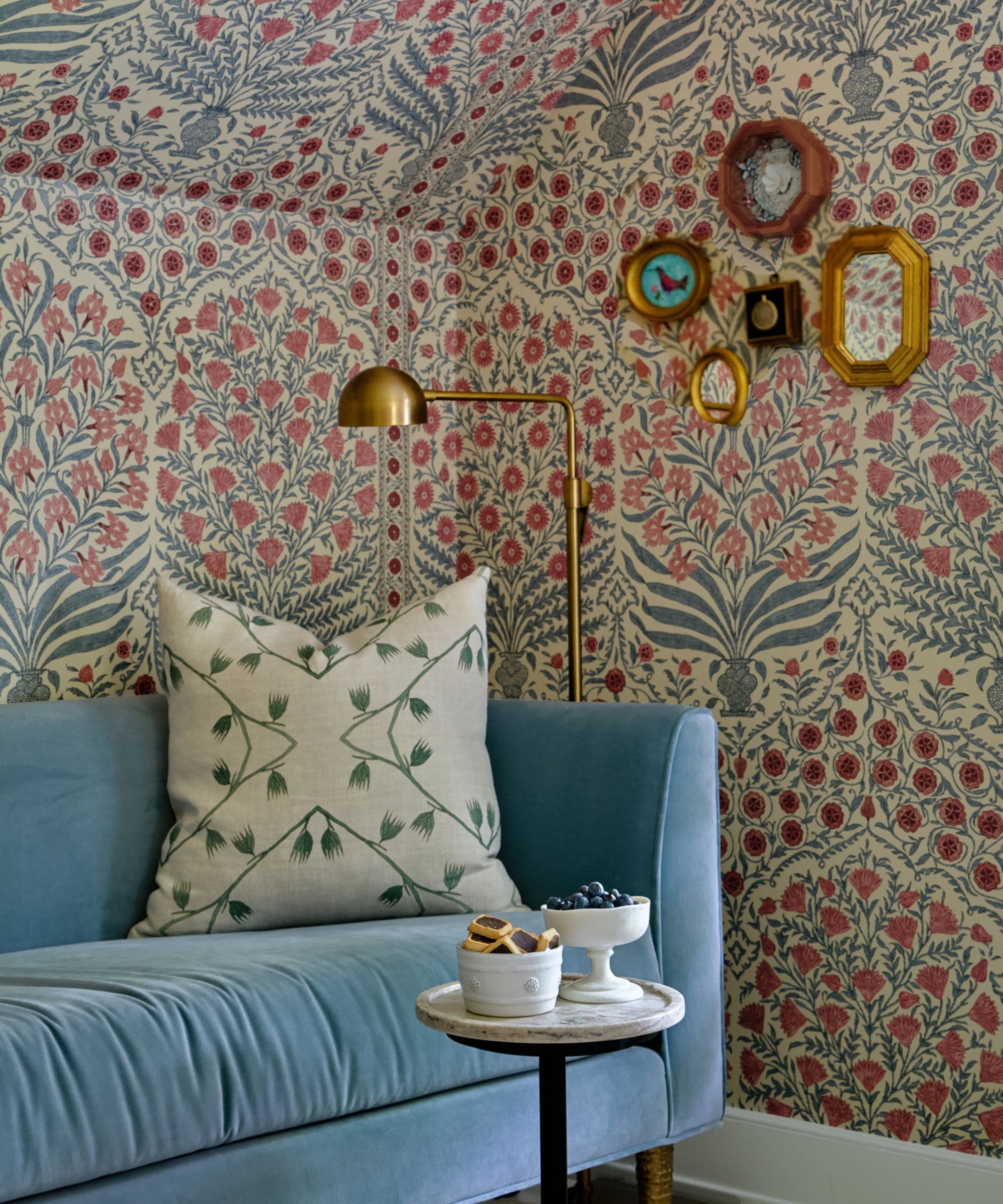Wallpapered bedroom with a cozy seating area featuring a pale blue velvet sofa, a small gallery wall with vintage prints, and a marble side table with snacks, and a gold metal floor lamp.
