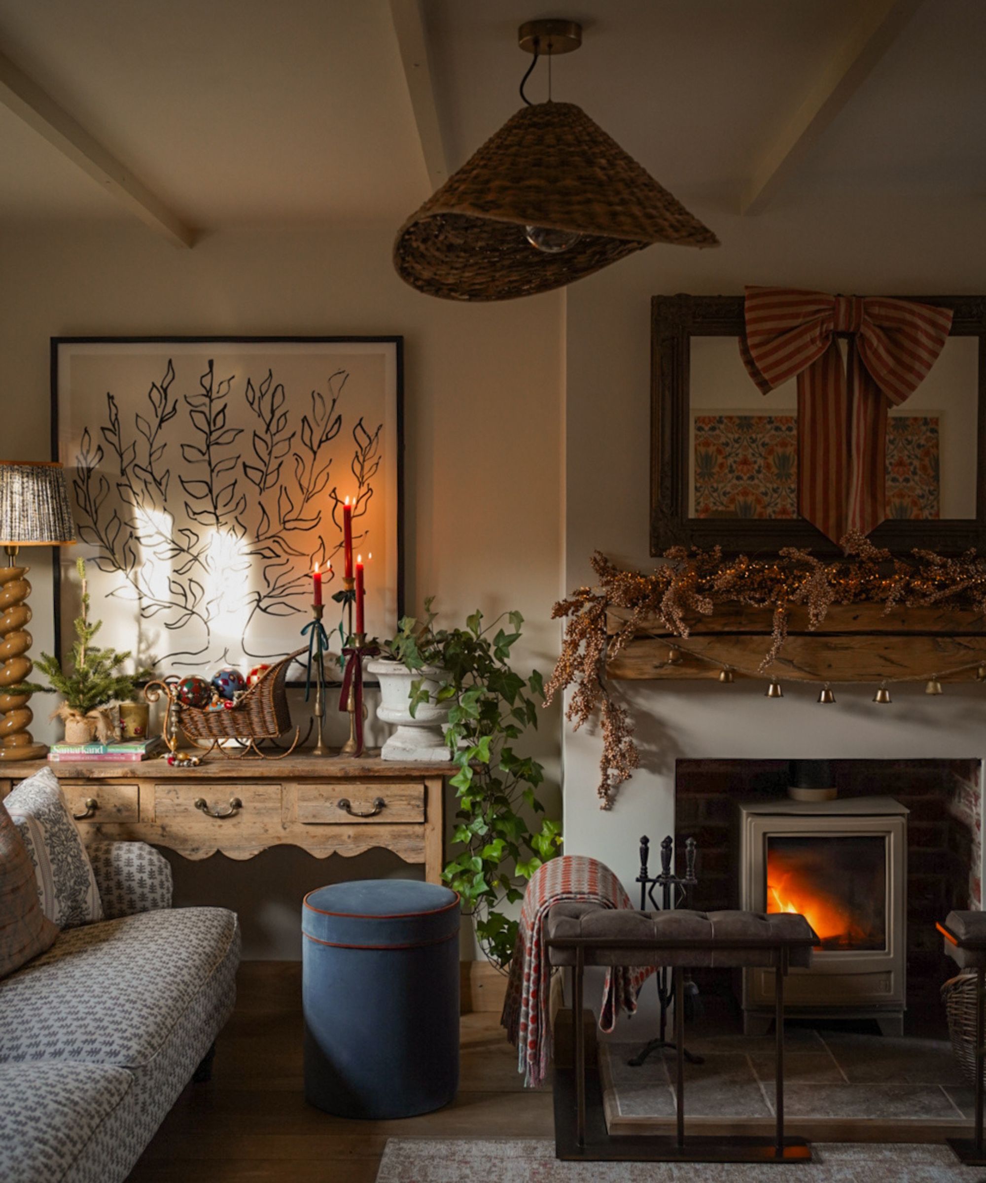 Rustic living room with a large wooden mantelpiece decorated with foliage garland, a log burner, a wooden console decorated with candles and wicker decorations, a sofa, and a fireplace fender.
