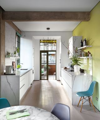 A modern white kitchen with statement kitchen wall decor ideas in lime green and a marble table with colorful blue chairs.