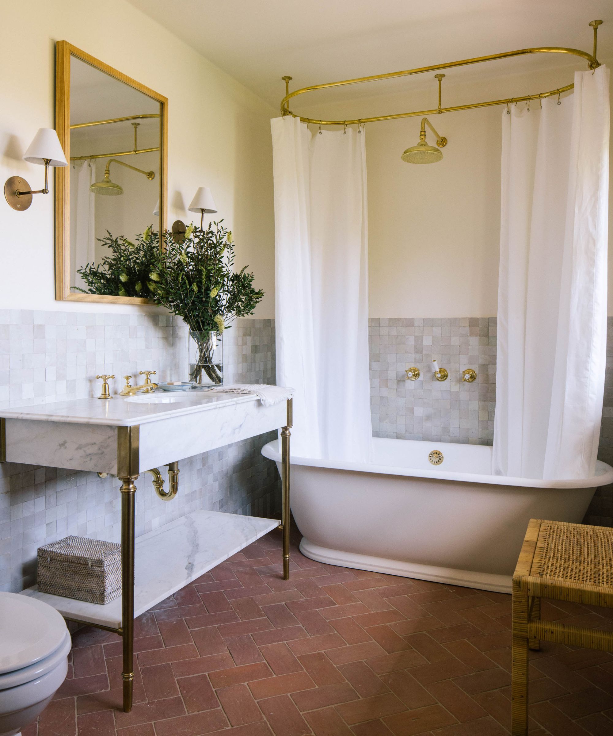 mediterrean style bathroom with terracotta floor tiles, neutral walls, zellige tiles at half height on the walls, a freestanding tub with brass hardware and white shower curtains with a marble vanity