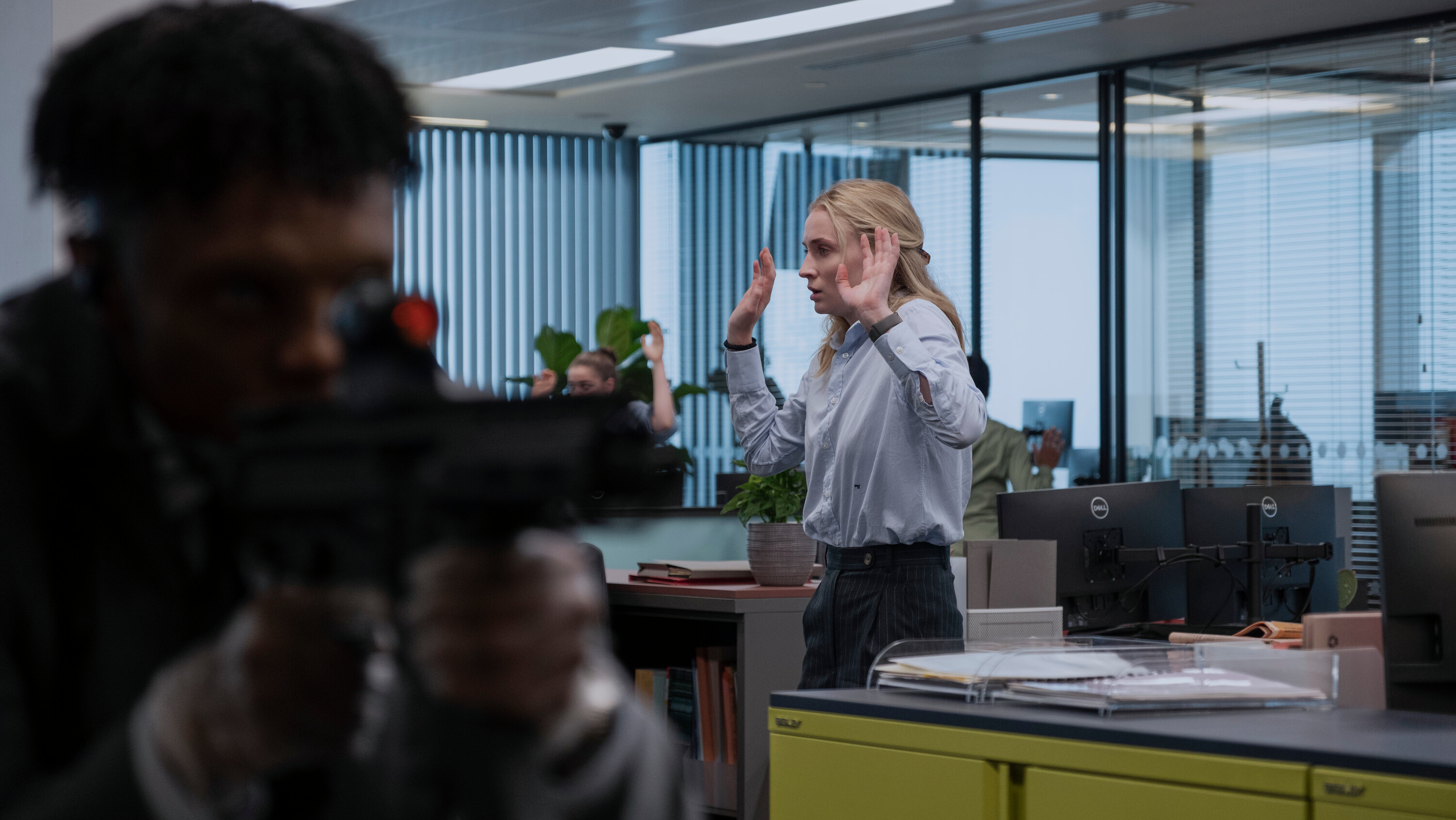L-R: A gun-wielding Yorkshire (Charles Mnene) walks through the Lochmill Capital office out of focus in the foreground, with Zara (Sophie Turner) surrendering in the background in a scene from "Steal" on Prime Video