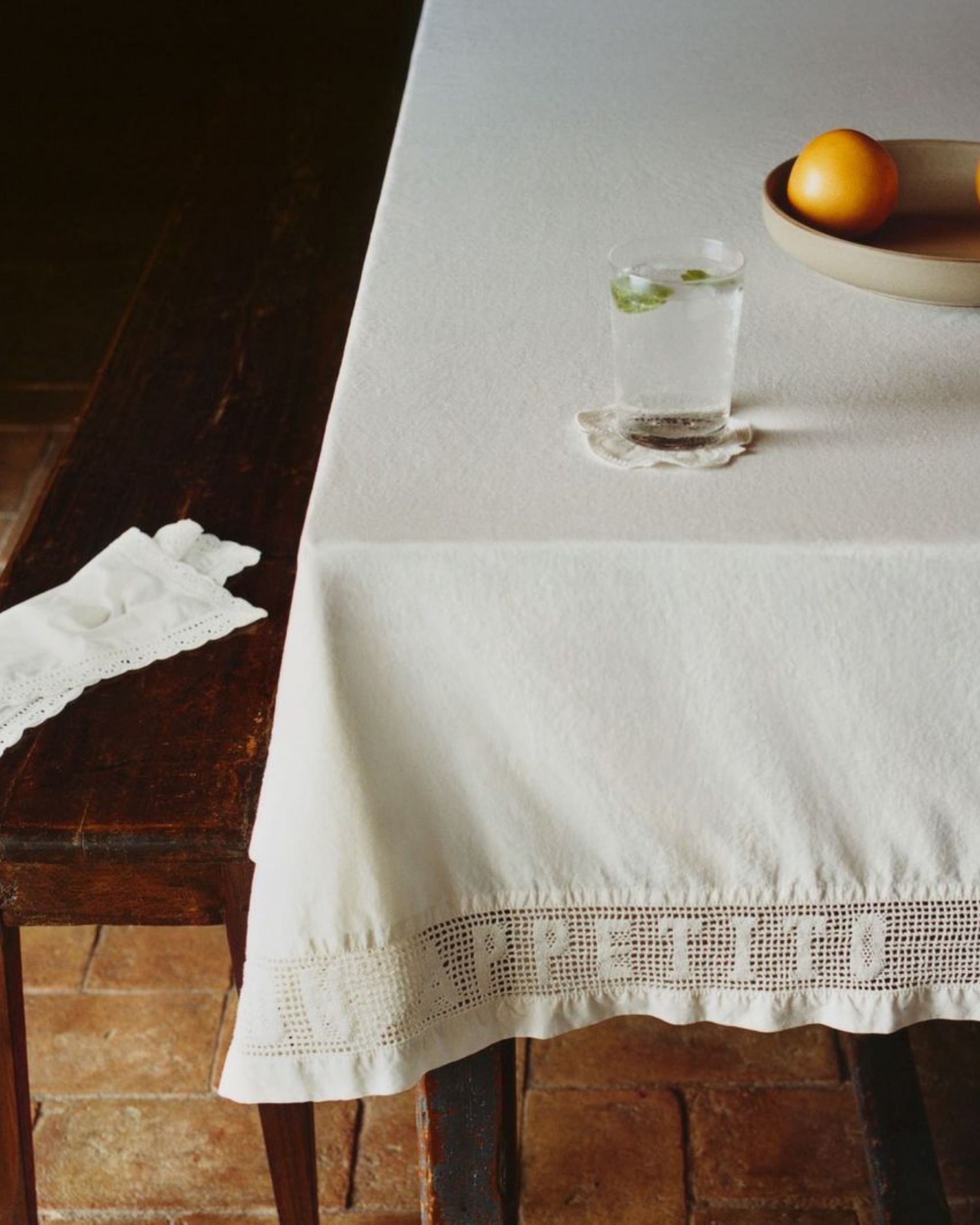 Image of a white tablecloth on a table with a wooden bench beside it and brick flooring.