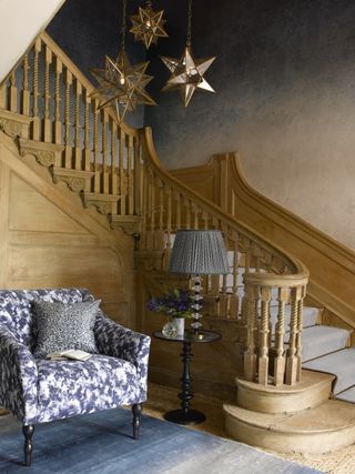 Hallway with blue patterned armchair, side table and lamp, blue rug and staircase with runner