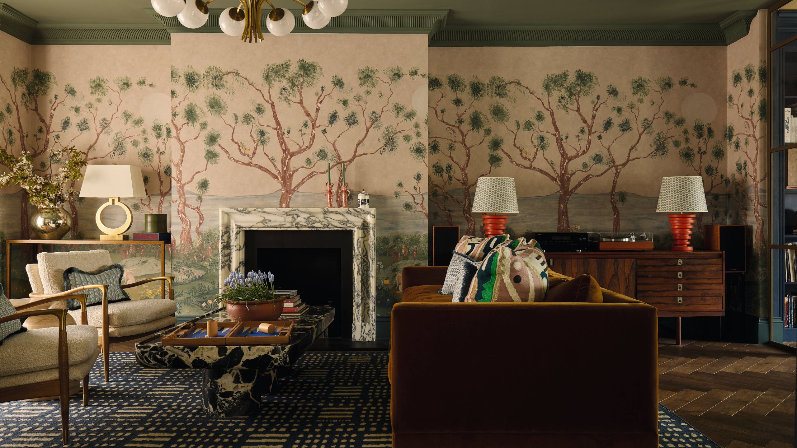 a large London living room with pink and tree motif mural wrapping the walls and chimney breast, a green painted ceiling, marble fireplace and eclectic furniture around a toffee coloured velvet couch