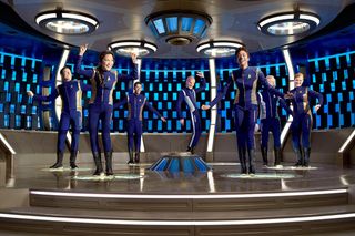 seven people in purple spaceflight uniforms stand on a futuristic-looking set.