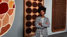 A young man dressed in a chequered blue and beige jacket, grey top and trousers stands in front of a series of colorful, tufted rugs hanging on a wall.