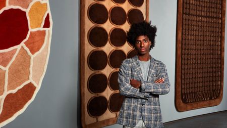 A young man dressed in a chequered blue and beige jacket, grey top and trousers stands in front of a series of colorful, tufted rugs hanging on a wall.