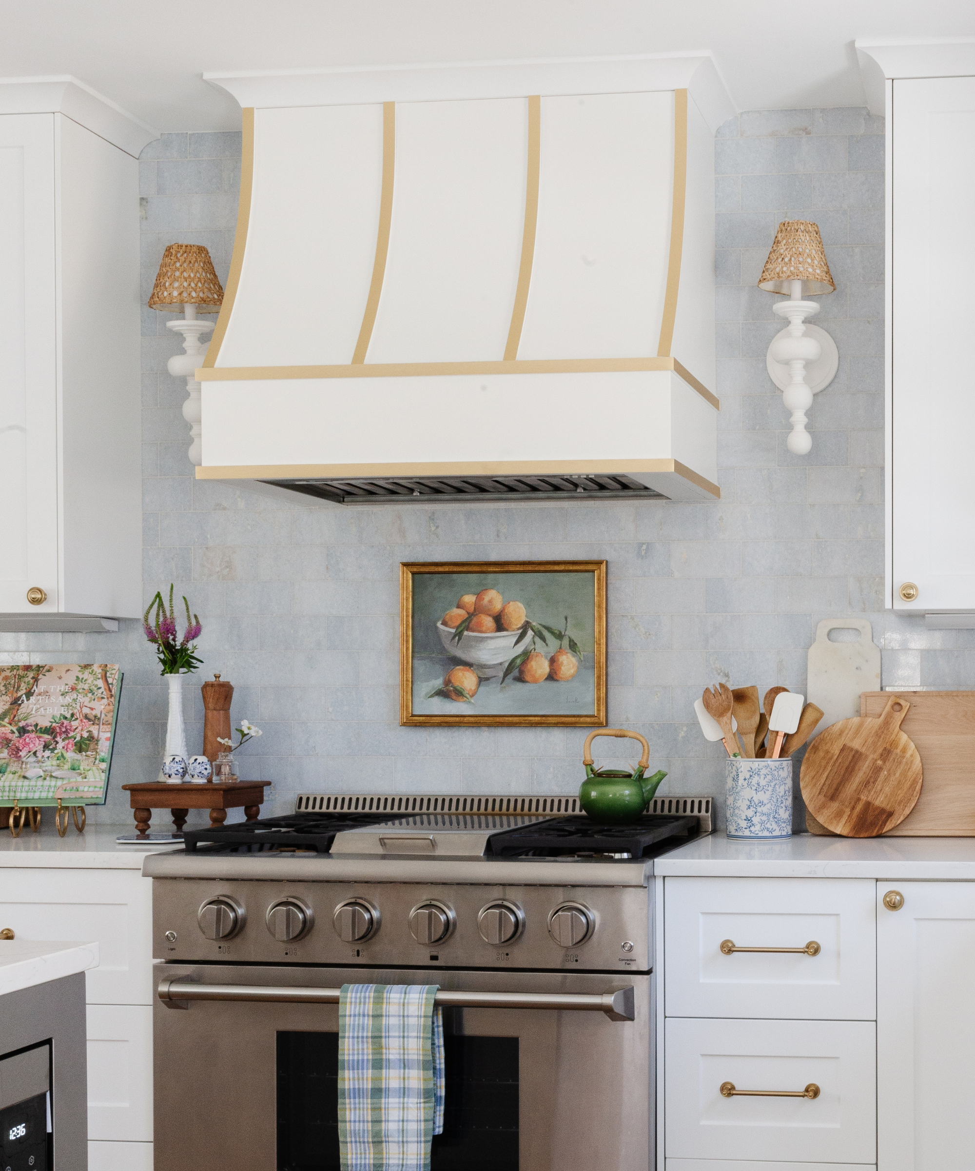 a bright kitchen with a light blue backsplash, white cabinetry, countertop utensils, a stainless steel stove, a white range hood, two wall sconces, and a painting of oranges behind the stove