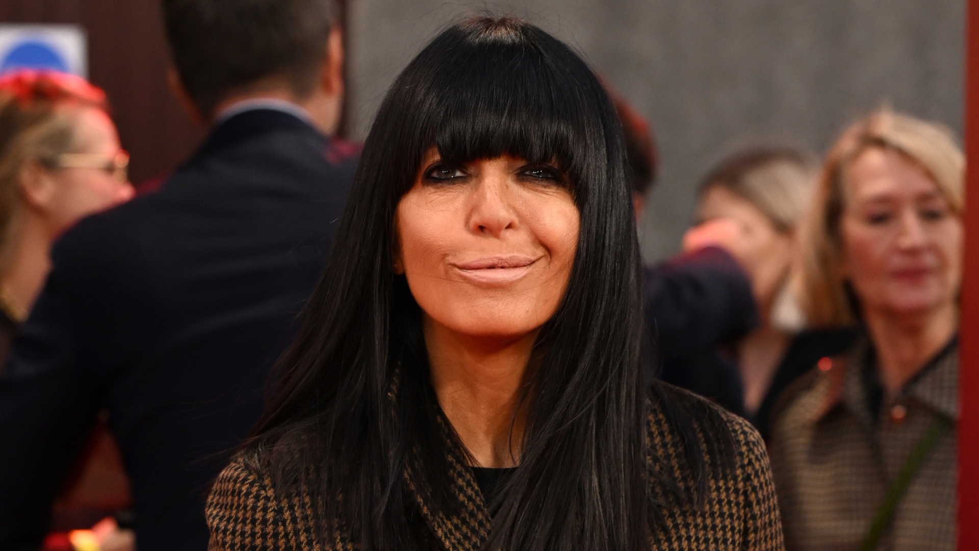 Image of Claudia Winkleman smiling with straight black long bob with full fringe and wearing a brown houndstooth blazer
