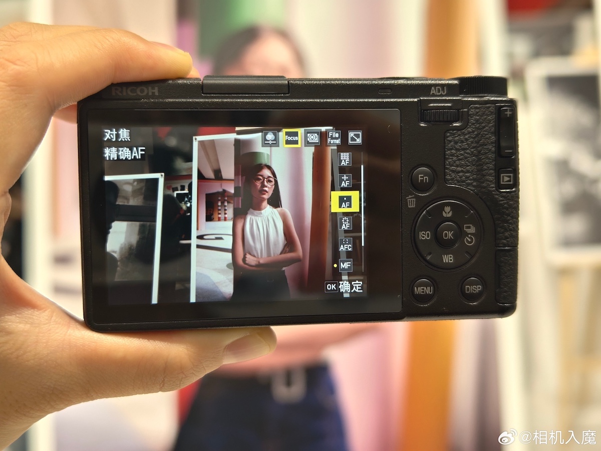 A hand holds a camera - Ricoh GR IV -displaying a photograph on its screen, showcasing a person standing against a colorful backdrop