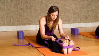 A woman with long brown hair performs a seated forward bend using a purple strap. She is on an orange yoga mat and there is a purple yoga block to the side.
