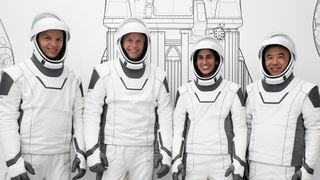 four astronauts in white spacesuits stand in a line. to their back is a diagram of a rocket on a white wall