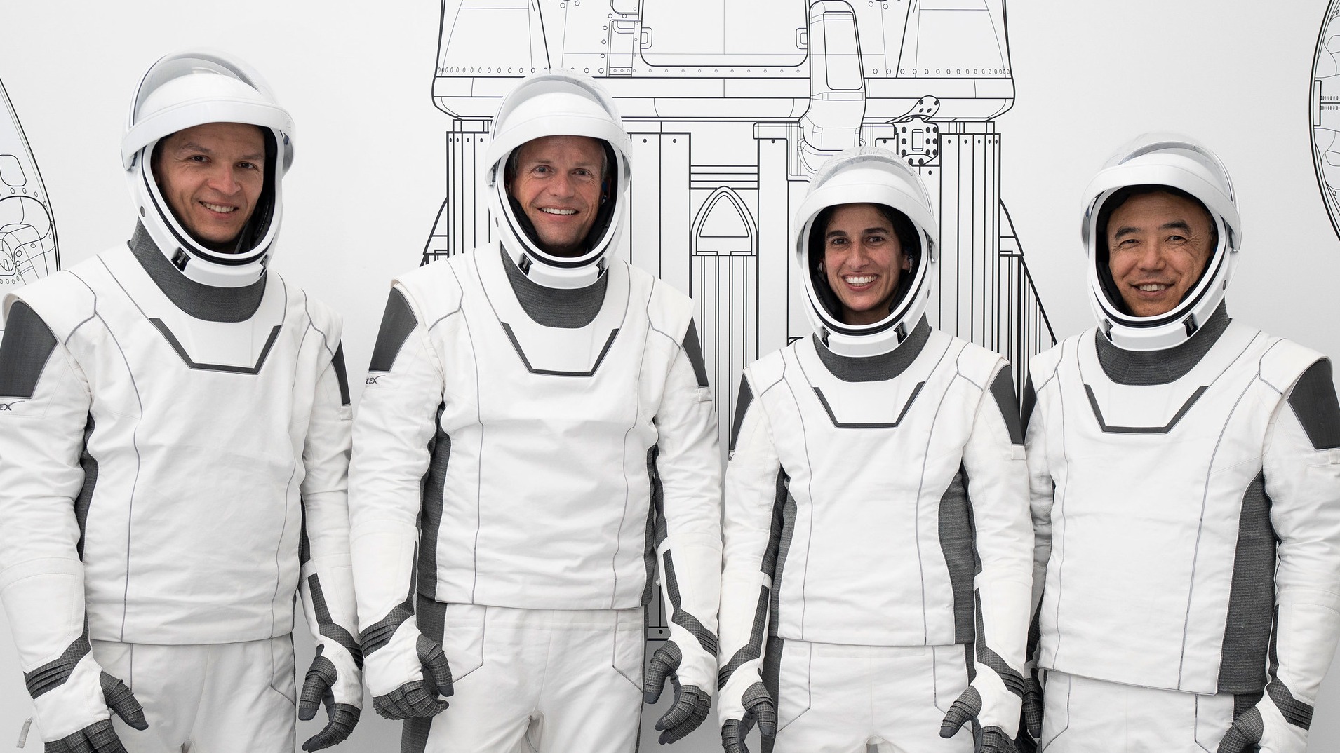 four astronauts standing in a row in spacesuits, smiling. a line drawing of a rocket is behind them