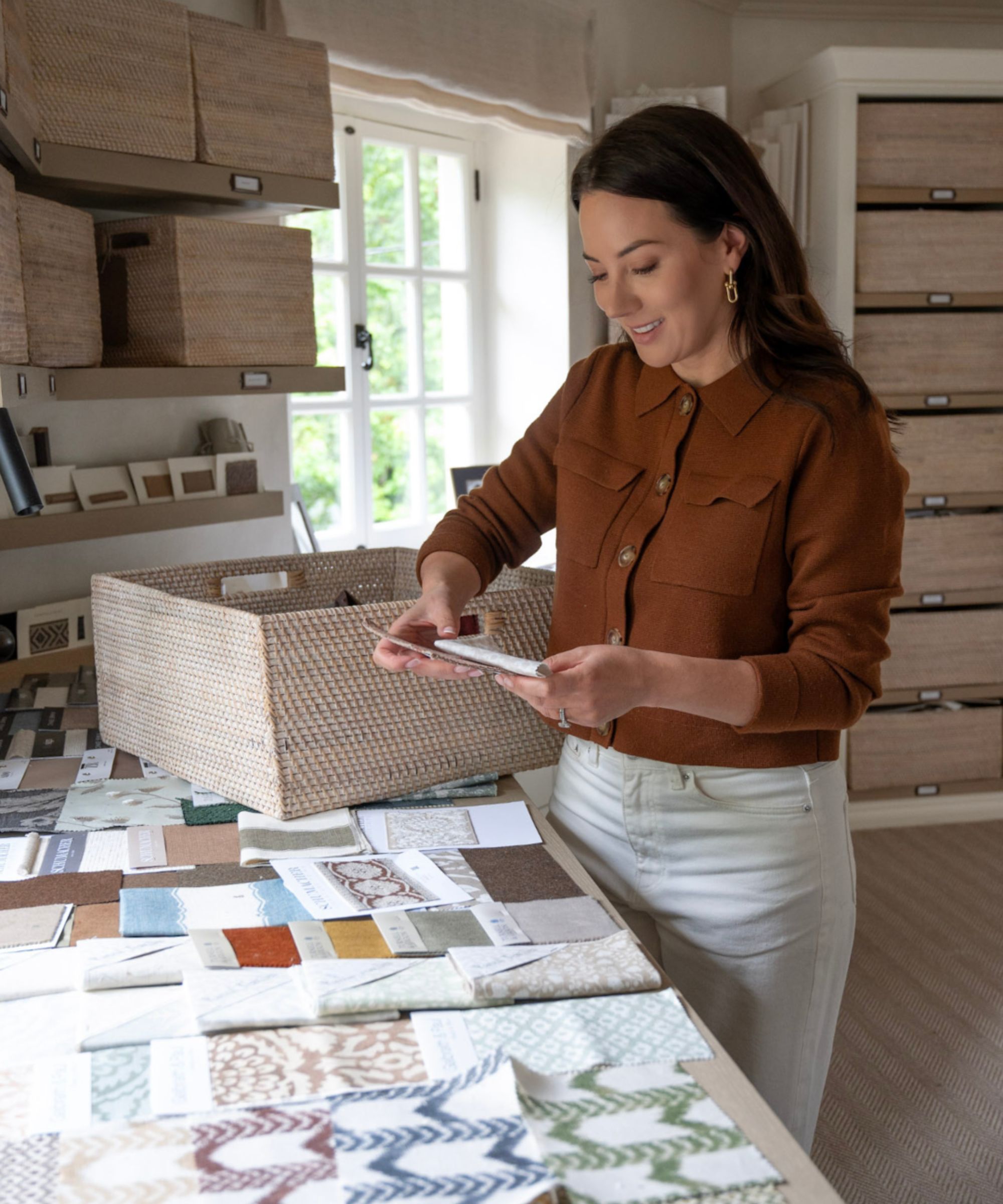 Sophie Paterson looking at fabric samples, laid out on a large worksurface, storage baskets and shelves in background