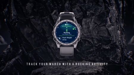 A video still of the Garmin Tactix 8 unveiling, showing a Rucking activity on the start screen with "Pack Weight: 25 lbs." underneath. Under the watch, the words "track your march with a rucking activity" are superimposed over a rocky background.