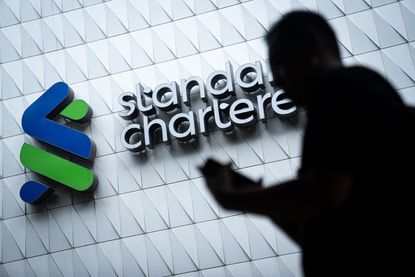Photograph of Standard Chartered sign and logo with a human shadow in the foreground. 