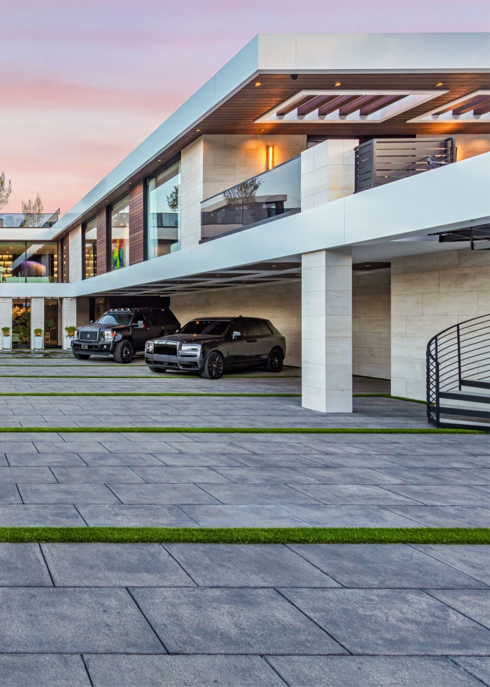 A large front yard with two cars part outside of a large penthouse in the US. There are two levels and in the front garden are stones flooring laid out with turn between the stone creating the illusion of levels and making the space feel bigger
