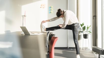 A woman in an office performs a leg stretch. She is in jeans, a blouse and sneakers. Her left leg is on the floor and her right leg is on a desk straight in front of her. She leans over her right leg and holds her toes with her right hand. Behind her we see a whiteboard with post-its and a plant. In front of her are a desk, desk chairs and a laptop. 