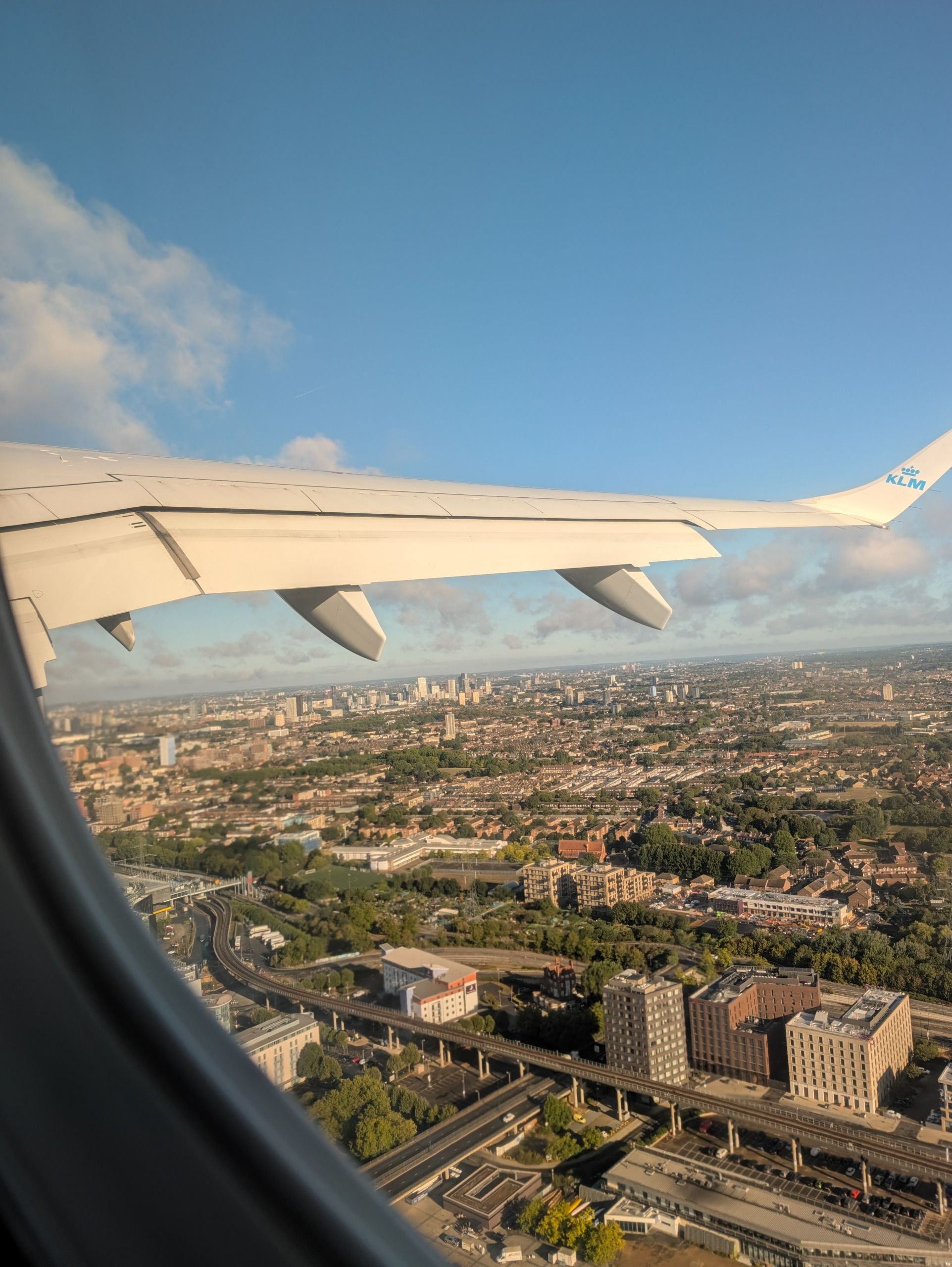View from an airplane window showing a KLM aircraft wing above a sprawling city in warm evening light.