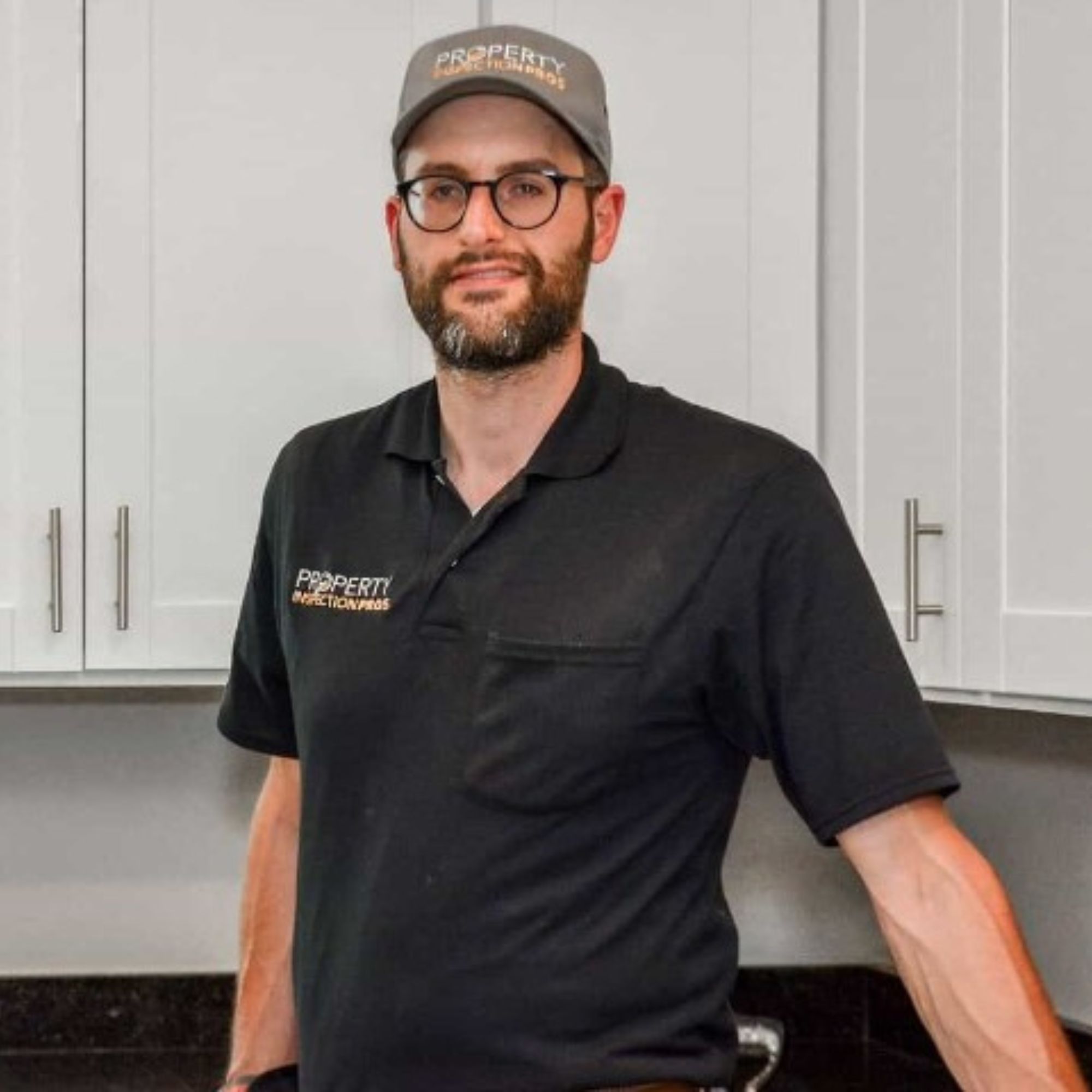 Sol Kruk, a white man in a black shirt, glasses, and cap, standing in a kitchen with white cupboards
