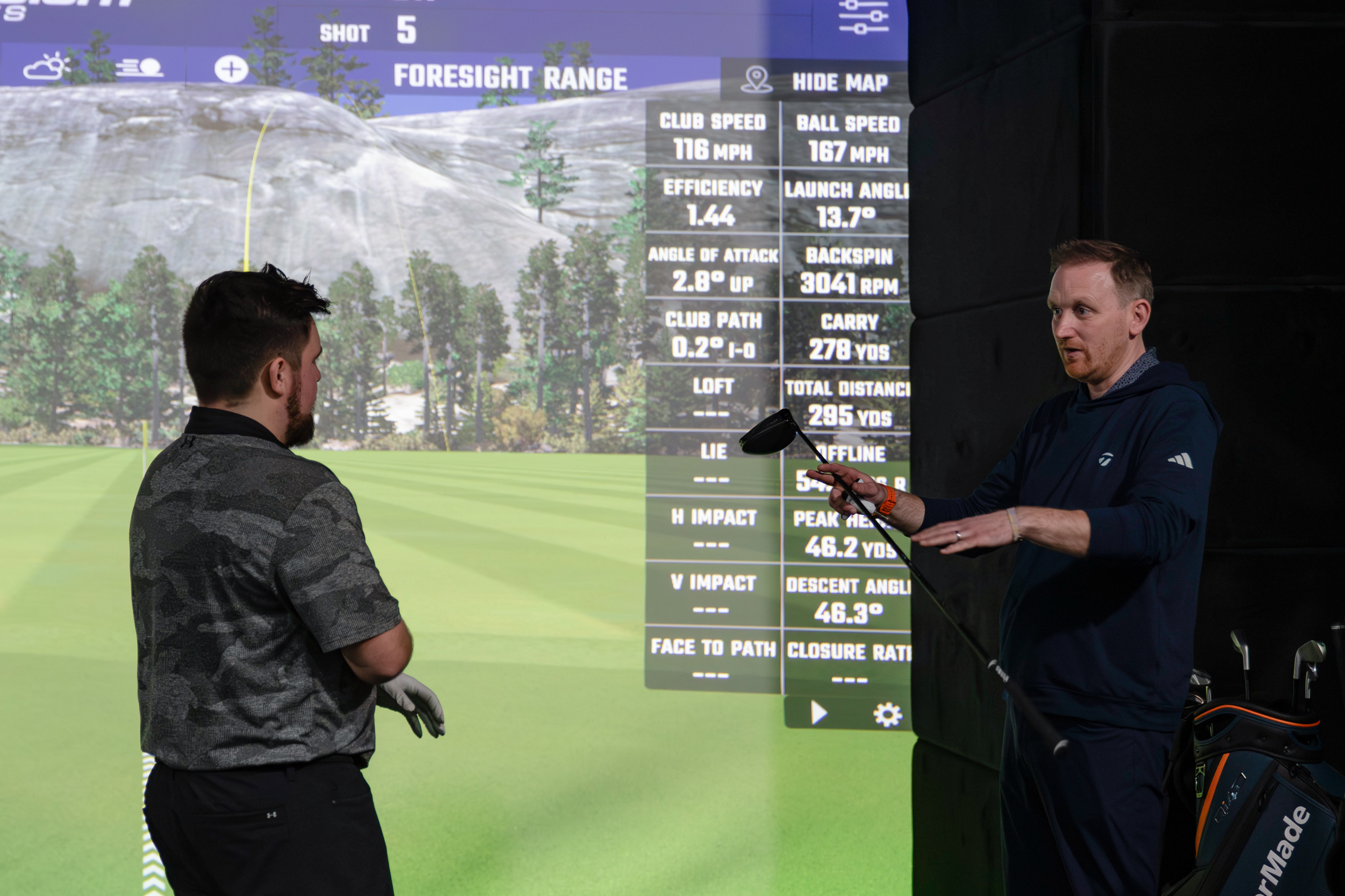 A fitter speaks to a golfer in front of a screen, holding a driver
