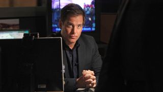 Tony DiNozzo seated at his desk and speaking with someone standing in front of him on NCIS