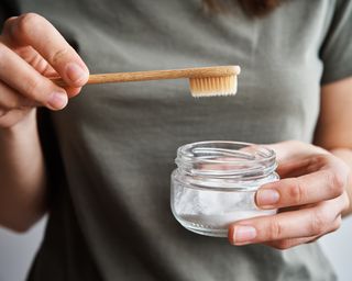 a woman holding a jar of toothpaste and a toothbrush - GettyImages-1317409509