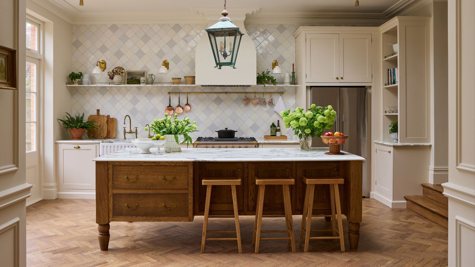 A wide shot of an elegant kitchen with herringbone wood floors, featuring a white fluted farmhouse sink, brass hardware, and a marble-topped oak island. Natural light streams in from a large window on the left, illuminating the soft cream and grey diamond tiles
