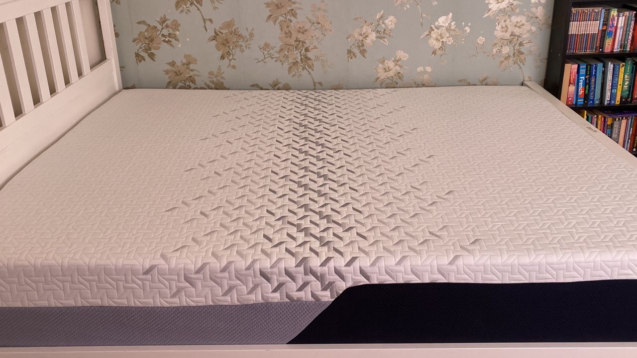 A queen size Nectar Premier Memory Foam Mattress on a white bedframe with a headboard in our tester's blue-and-gold bedroom. A bookcase can be seen in the top right-hand corner of the image.