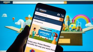 Person, die ein Smartphone hält und auf die Amazon-App schaut, mit einem Monitor dahinter, der die Amazon-Website zeigt