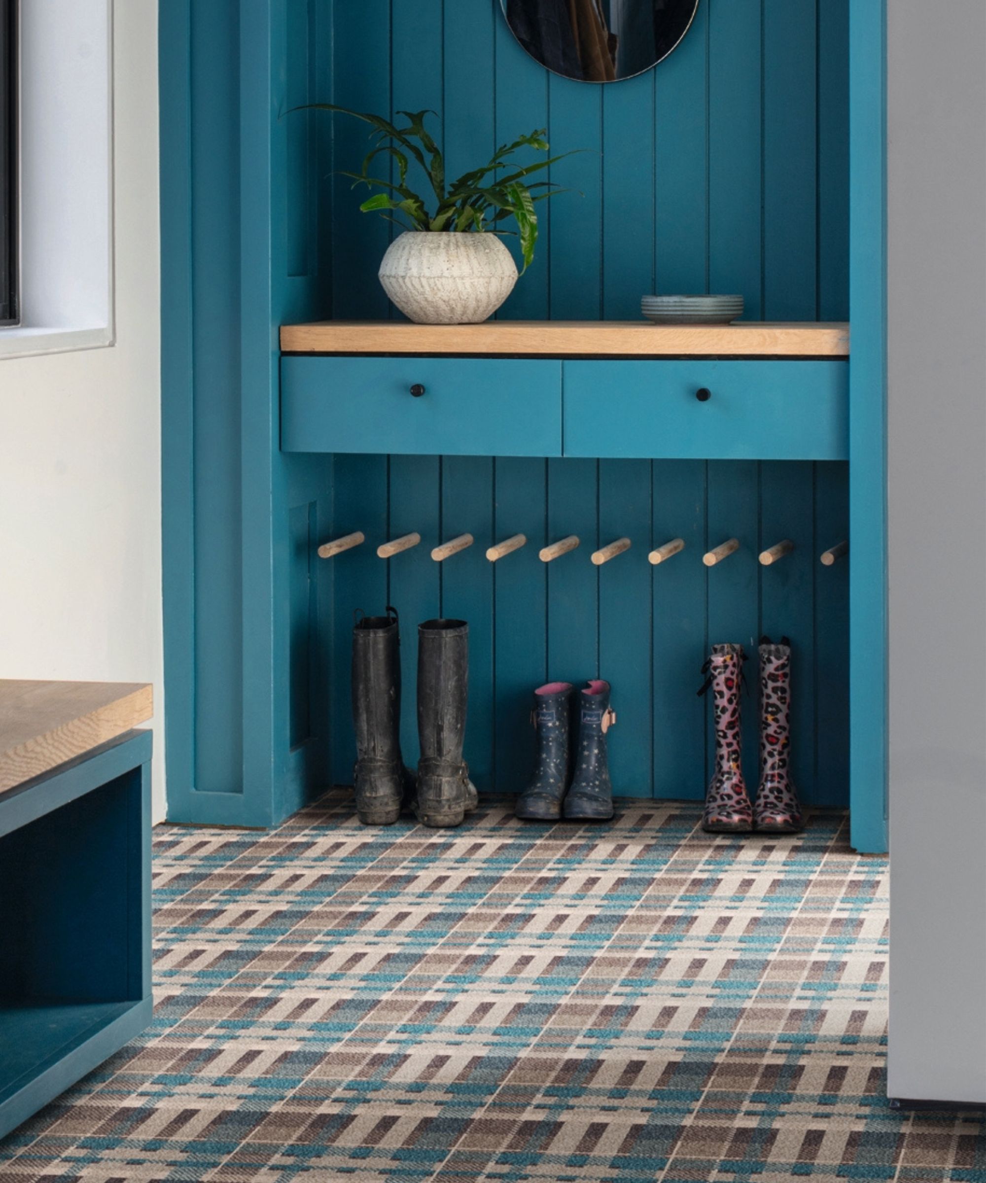 A vibrant teal painted built-in hallway boot room with tongue-and-groove panelling, an oak shelf with two small drawers and a potted plant above, and a row of wooden dowel pegs below. Three pairs of wellington boots in varying sizes stand on a geometric patterned tile floor.