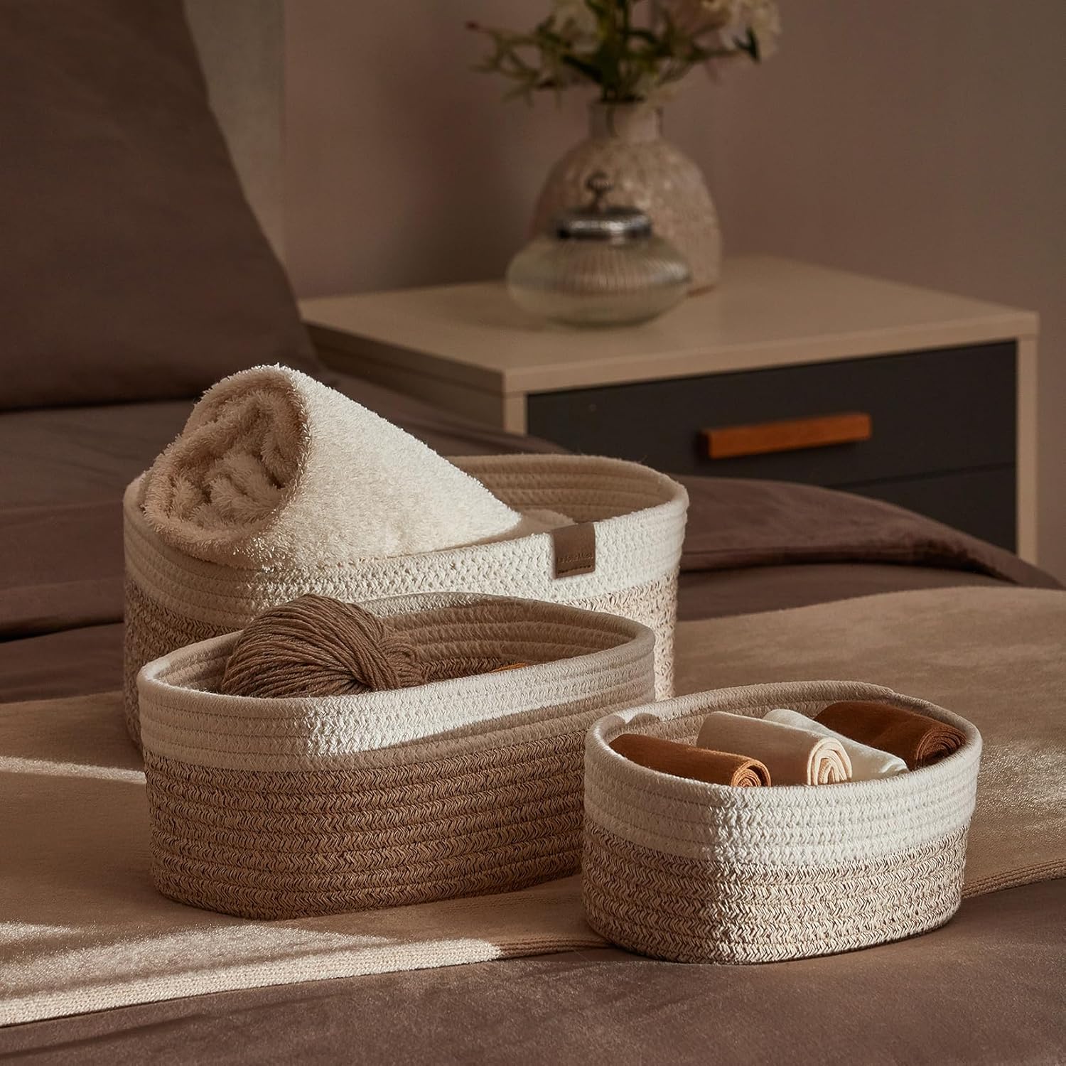 Three soft, woven storage bins with towels and other textiles in them. They are sitting on a made bed with neutral bedding.