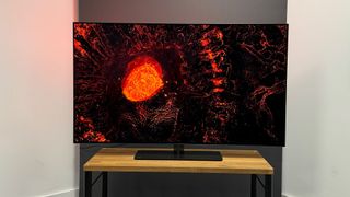 The OLED808 on a wooden stand against a white and grey background. A black and red image featuring a large pool of lava is on the screen.