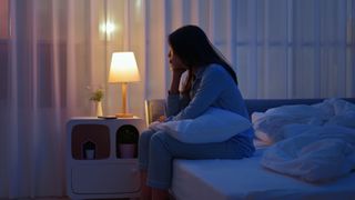 A young woman sits at the edge of her bed in a dimly lit bedroom as she can't sleep due to worry.